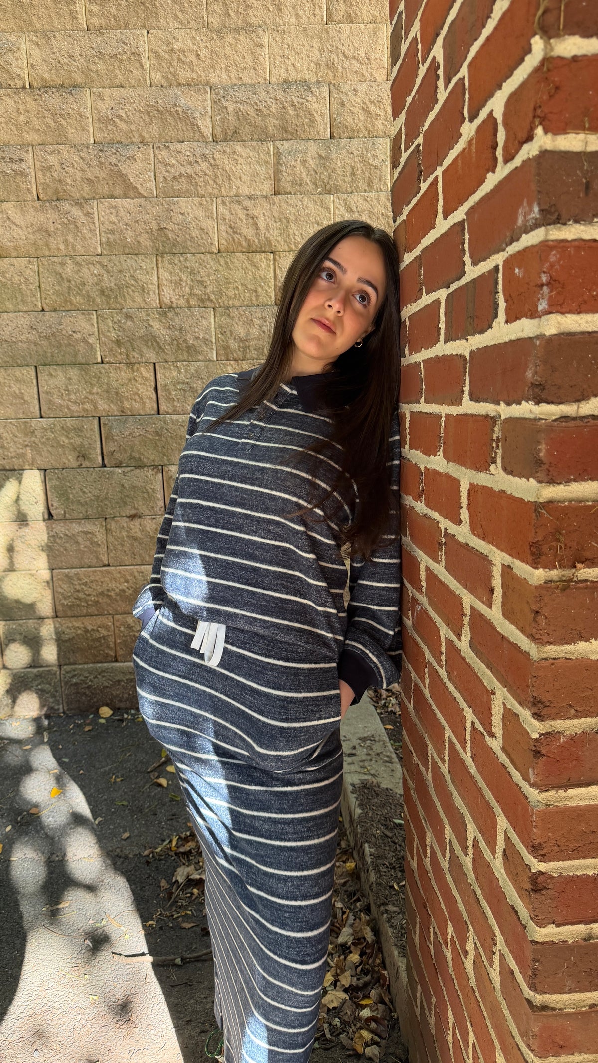 A woman wearing a matching navy and white striped lounge set, consisting of a long-sleeve top and pants, standing outdoors between a beige stone wall and a red brick wall.